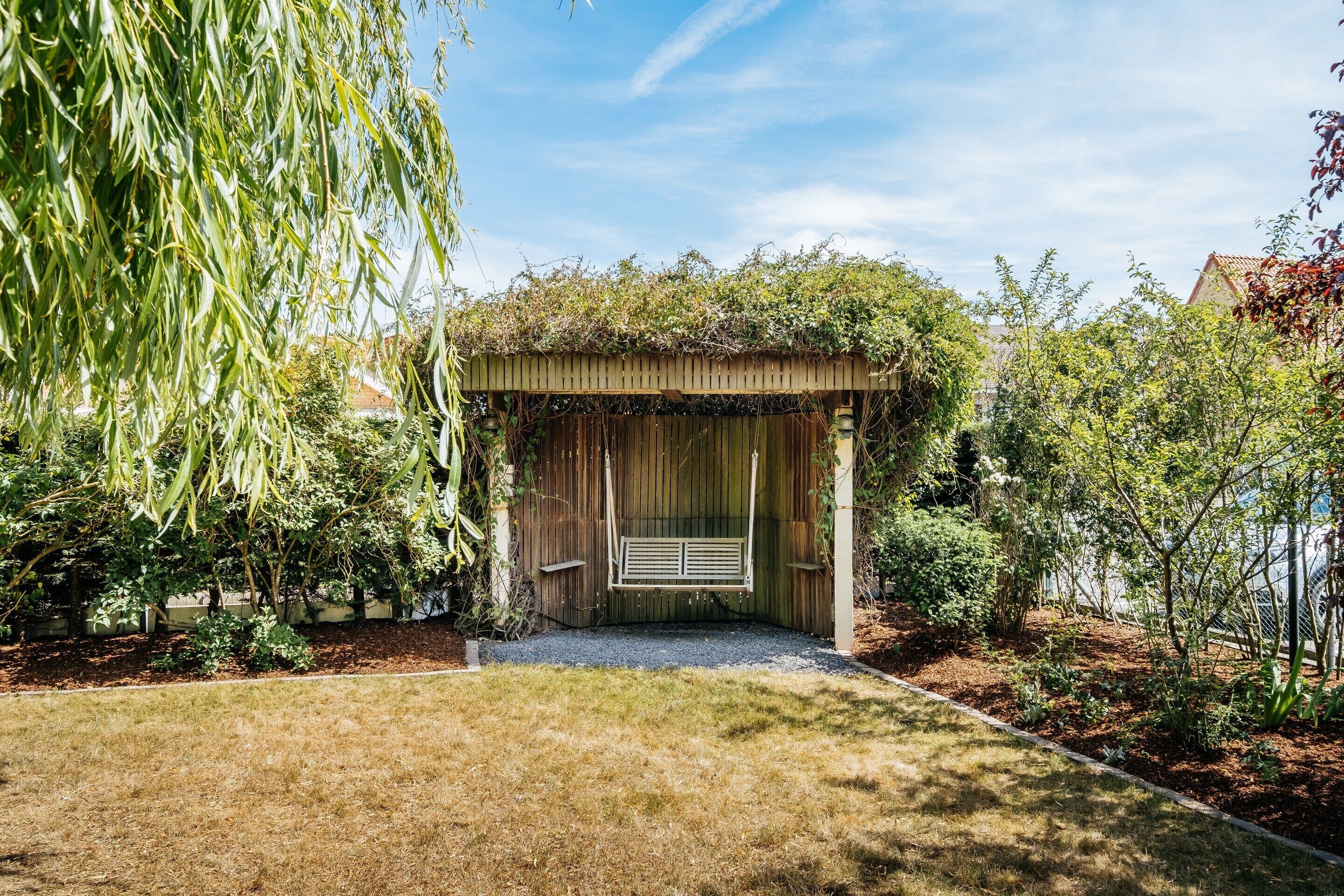 A wooden pergola with a swing, surrounded by lush greenery and a gravel path.