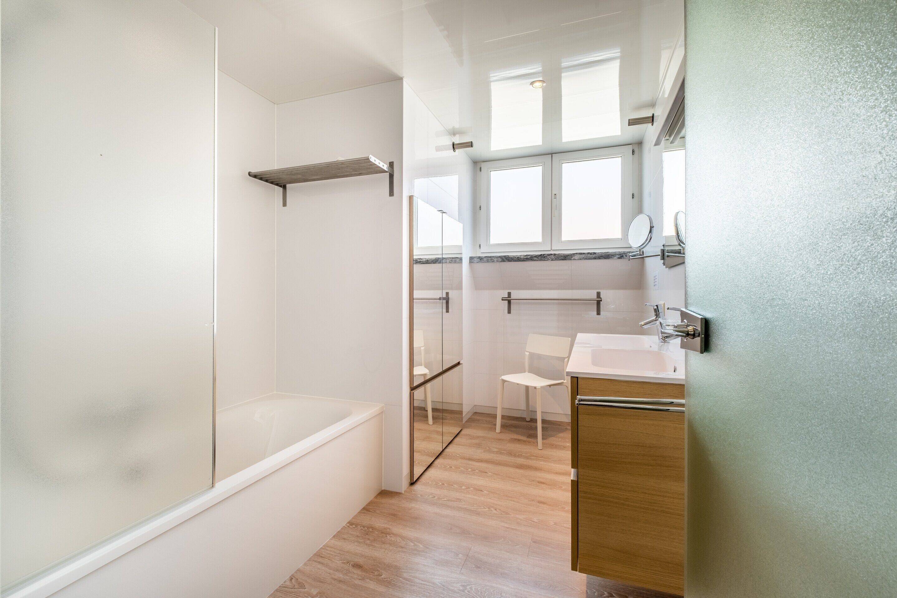 A modern bathroom with a glass shower enclosure, a wooden vanity, and a white chair.