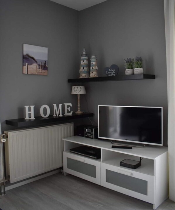 A modern living room with a grey wall, a white entertainment unit, a flat-screen TV, and decorative items including a 'HOME' sign, a lamp, and a shelf with figurines and plants. - Holiday Apartment Mosselbank 95 (Ouddorp)