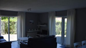 A living room with a sofa, a coffee table, and a TV. There is a sliding glass door leading to a balcony with greenery outside. - Holiday Apartment Mosselbank 95 (Ouddorp)