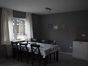 Een eetruimte met een tafel gedekt voor zes personen, donker houten stoelen, een raam met gordijnen en een klein kastje aan de rechterkant.