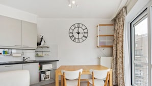 A modern kitchen with white cabinets, a wooden dining table with four chairs, a large wall clock, and a window with curtains. - Fiesole 201 Very Centrally Located in Koksijde (Koksijde)