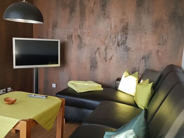 Una sala de estar moderna con un sofá de cuero negro, un televisor de pantalla plana montado en la pared, una mesa de madera con mantel verde y una lámpara.