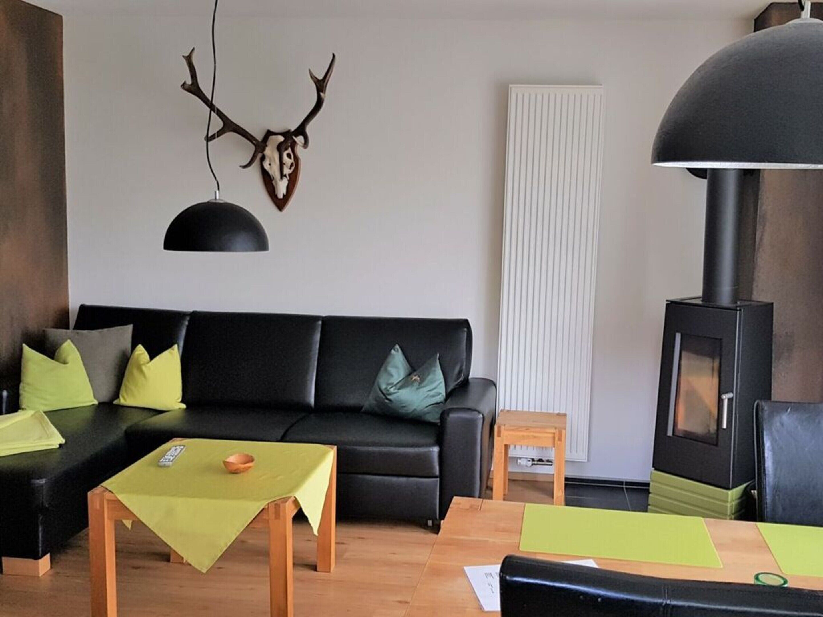 Un salon moderne avec un canapé noir, une petite table en bois, une suspension noire, un crâne de cerf accroché au mur et un poêle à bois.