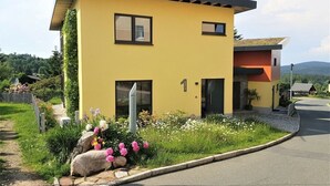 A modern yellow building with a flat roof, a small attached structure with a red roof, and a well-maintained garden with flowers and a rock feature. - Gutsweg One - Apartment 3 (Eibenstock)