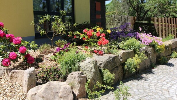 A modern building with a yellow facade and large windows, surrounded by a landscaped garden with various colorful flowers and a stone pathway. - Gutsweg One - Apartment 3 (Eibenstock)