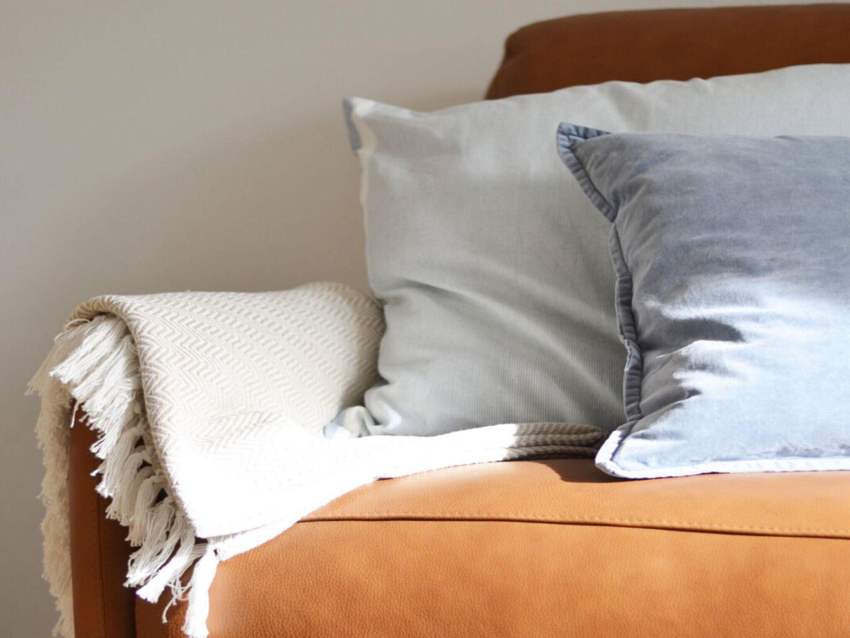 A room with a brown leather sofa, a white throw blanket, and two pillows, one light blue and one white.