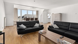 Amplia sala de estar con dos sofás negros, una mesa de centro de madera y vistas de la ciudad a través de grandes ventanales.
