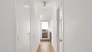 Un pasillo estrecho con paredes blancas y suelo de madera, que conduce a una puerta desde donde se ve una vista de la ciudad a través de una ventana.