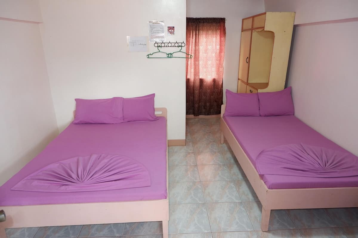 A room with two beds, each with a purple mattress and matching pillows. There is a wooden wardrobe against the wall and a small shelf with a mirror.