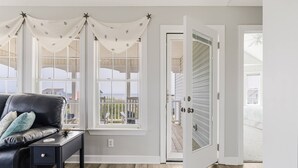 A room with a black leather sofa, a small side table, and a window with white curtains. There is a door leading to an outdoor area. - Gorgeous 2nd Row Home with Amazing Ocean Views, 2 Large Decks, Dog Friendly, Sleeps 12 (Oak Island)