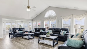 A spacious living room with a high ceiling, large windows, and a view of the outside. There are multiple black leather sofas and a coffee table. - Gorgeous 2nd Row Home with Amazing Ocean Views, 2 Large Decks, Dog Friendly, Sleeps 12 (Oak Island)