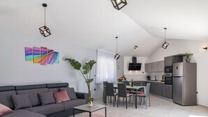 Eine moderne Wohnung mit grauem Sofa, einem Esstisch mit Stühlen, einer Küche mit Kühlschrank sowie einem Wohnbereich mit Pflanze und Kunstwerken.