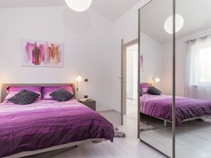 Une chambre à coucher moderne avec un grand miroir, un lit avec une literie violette et une table de chevet avec une lampe.