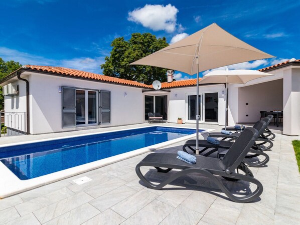 Une piscine entourée de chaises longues et agrémentée d’un parasol, le tout adossé à un bâtiment blanc doté d’un toit en tuiles.