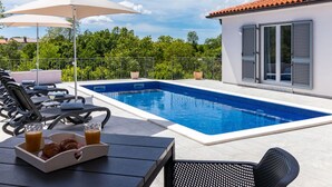 Ein Swimmingpool mit Liegestühlen und Sonnenschirmen, ein Esstisch mit Getränken und Gebäck, und ein Gebäude mit einem Ziegeldach und Fensterläden.