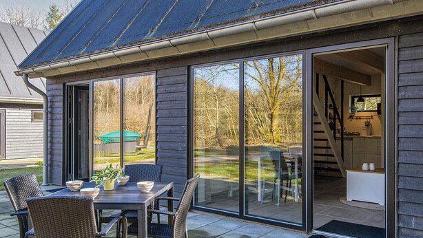 A modern house with a patio area featuring a dining table and chairs, large sliding glass doors leading to a kitchen, and a view of the surrounding landscape. - 5 Star Holiday Home in Hovborg (Hovborg)
