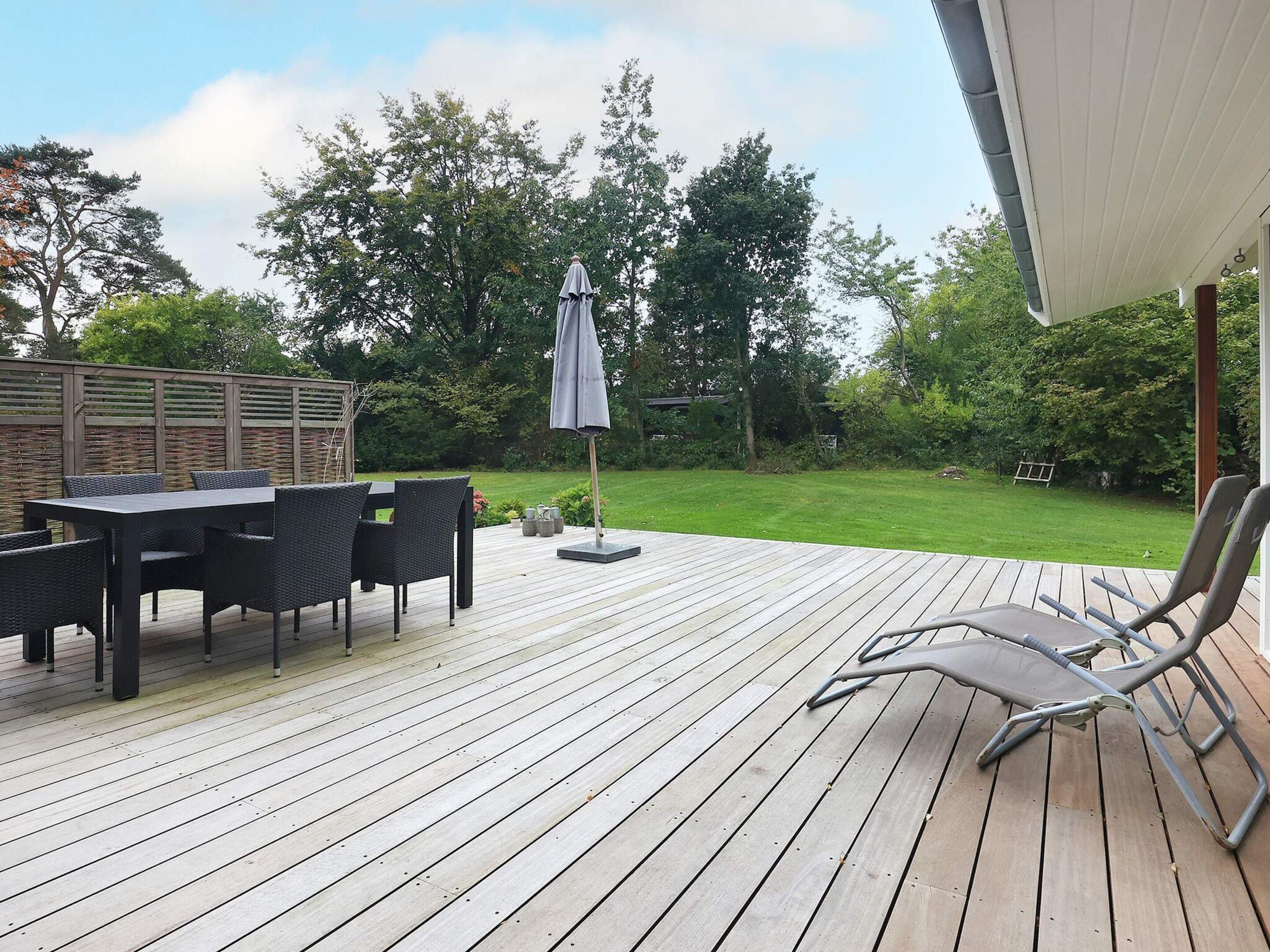 Una terraza de madera con mesa y sillas, un área cubierta y vista a un jardín con árboles.