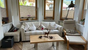 A cozy living room with a sofa, a coffee table, and a chair. There are windows with blinds, a lamp, and a small table with decorative items.