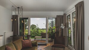 Un soggiorno moderno con un divano verde, un tavolino da caffè e una zona pranzo con tavolo e sedie. Ampie finestre offrono vista su un giardino e un edificio in lontananza.