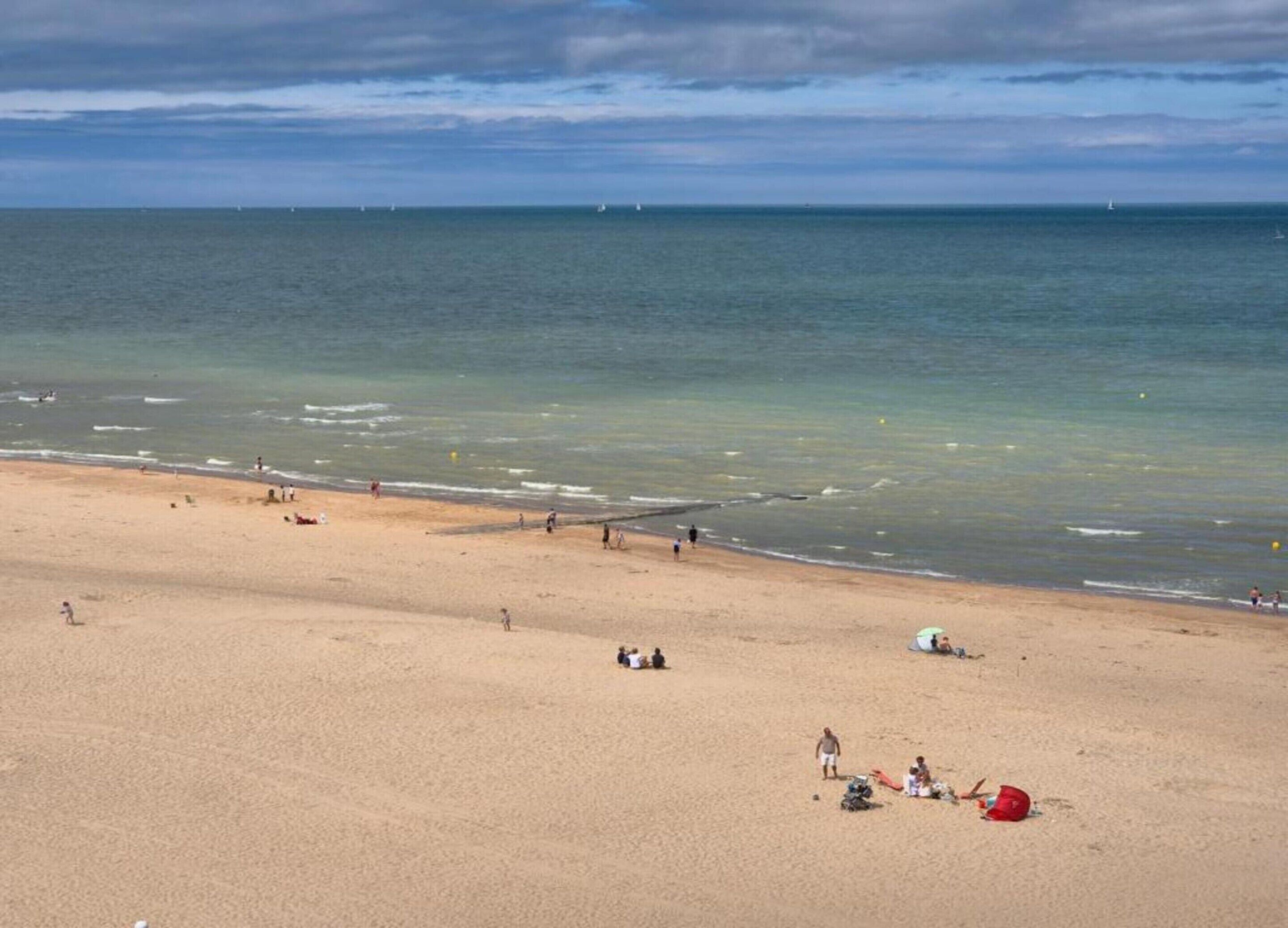 Una amplia playa de arena con un mar tranquilo, barcos a lo lejos y gente disfrutando de la orilla.