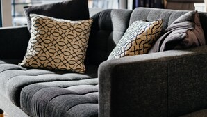 Ein graues Sofa mit gemusterten Kissen in einem modernen Zimmer mit Blick auf ein Backsteingebäude durch ein Fenster.