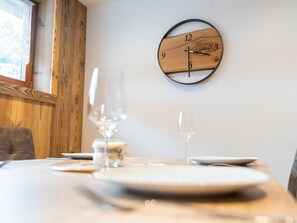 A dining table with plates, glasses, and cutlery, a wooden wall clock, and a window with a lace curtain. - Comfortable Apartment in Salzburger Land (Neukirchen am Grossvenediger)