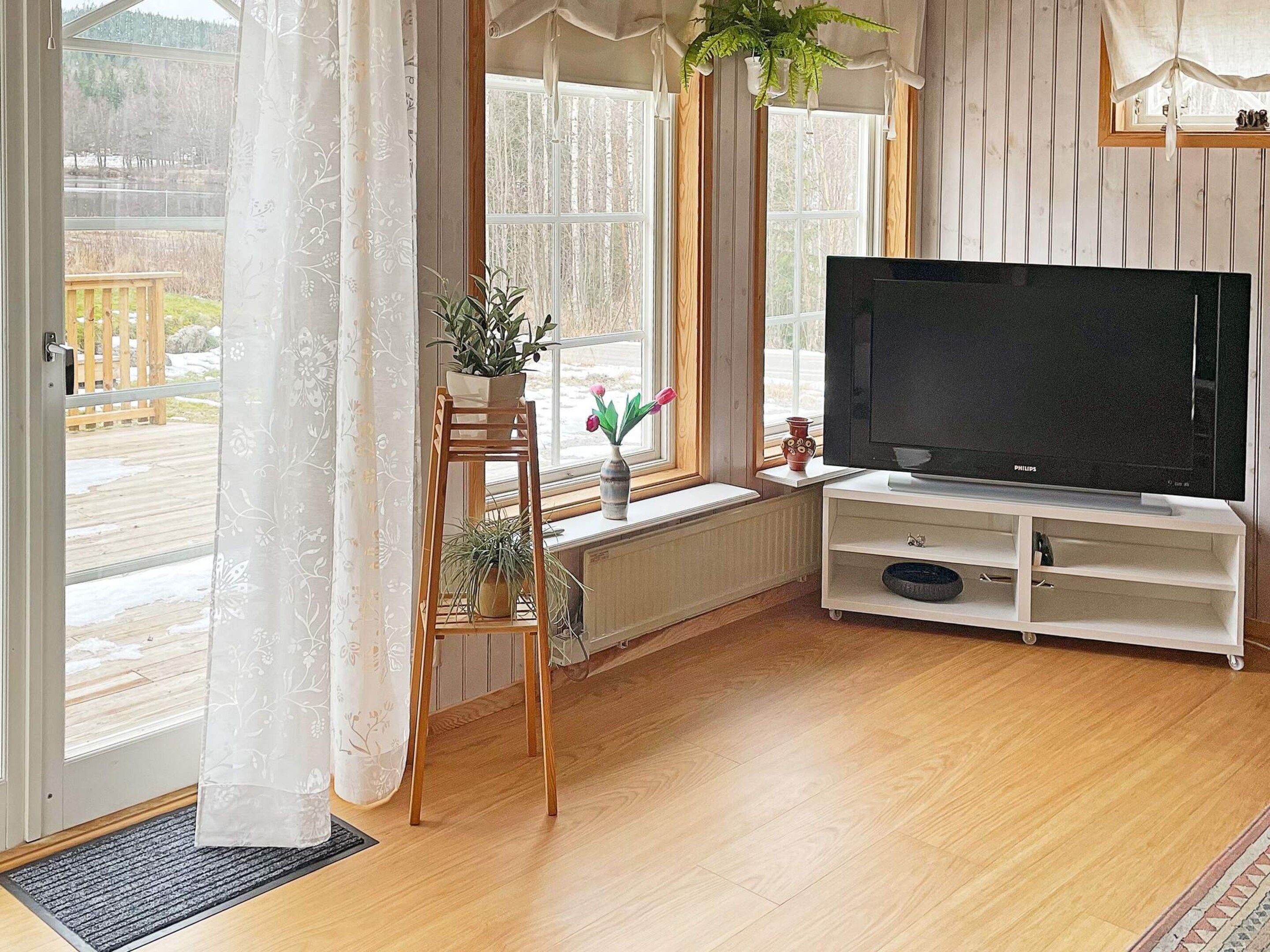 A living room with a television on a stand, a wooden floor, a window with curtains, and a view of a deck outside.