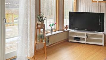 A living room with a television on a stand, a wooden floor, a window with curtains, and a view of a deck outside.