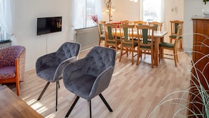 A modern living room with a wooden floor, a dining area with wooden chairs and a table, and a television mounted on the wall.