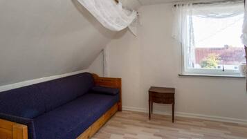 Ein Zimmer mit einer hölzernen Bank, einem kleinen Holztisch, einem Fenster mit weißen Vorhängen und einem Fußboden mit Fischgrätmuster.