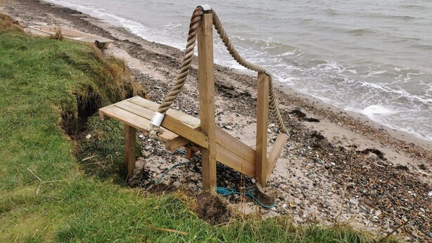 A wooden bench with a rope tied around it, located on a pebbled beach with the sea in the background.