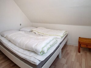 Un llit individual amb roba de llit blanca i un capçal de fusta en una habitació amb terres de fusta i parets blanques.