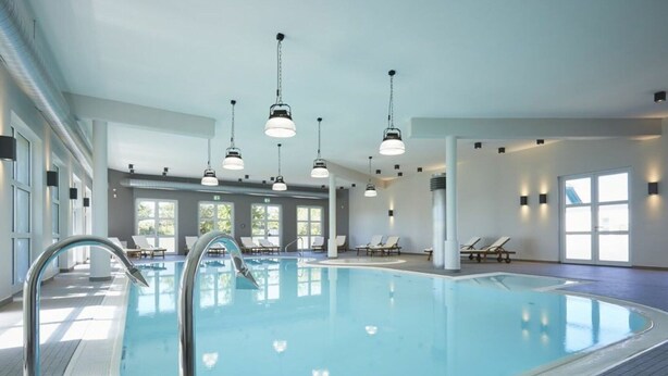 An indoor swimming pool with a clear water surface, surrounded by lounge chairs and sunbeds, with overhead lighting and large windows allowing natural light. - Lush Villa With Sauna and Fireplace (Dranske)