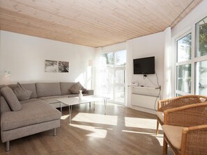 Un salon moderne avec un canapé d’angle, une table basse, des fauteuils en osier, un téléviseur à écran plat et de grandes fenêtres.