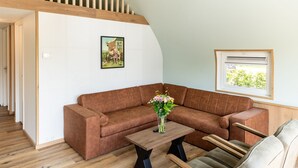 A modern living room with a brown sofa, a wooden coffee table with a vase of flowers, and a window with a view of greenery. - House With a Terrace on the Edge of the Forest (Baars)