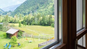 A view from a window overlooking a grassy field with a swing set, a wooden shed, and a mountain in the background.