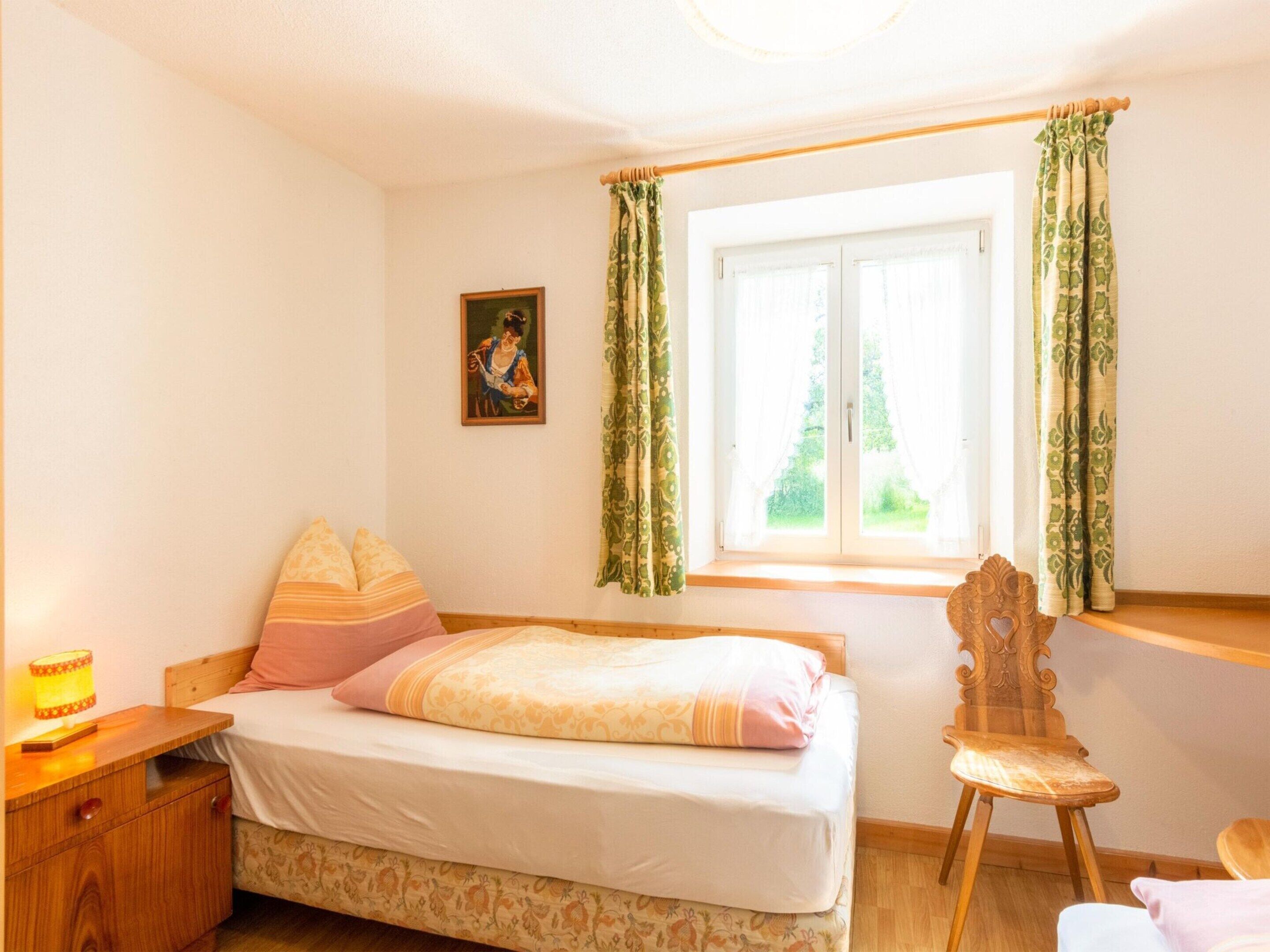 A bedroom with a bed, a nightstand, a chair, a window with curtains, and a painting on the wall.