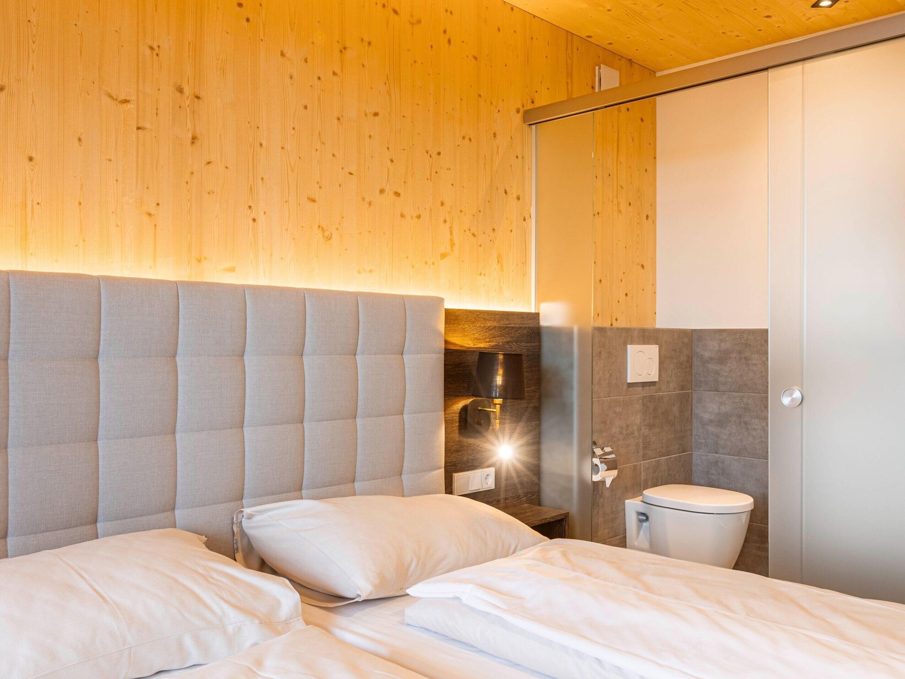 Ein modernes Hotelzimmer mit Holzboden, einem großen Bett mit grauem Kopfteil, einem Nachttisch mit Lampe und einer Toilette.