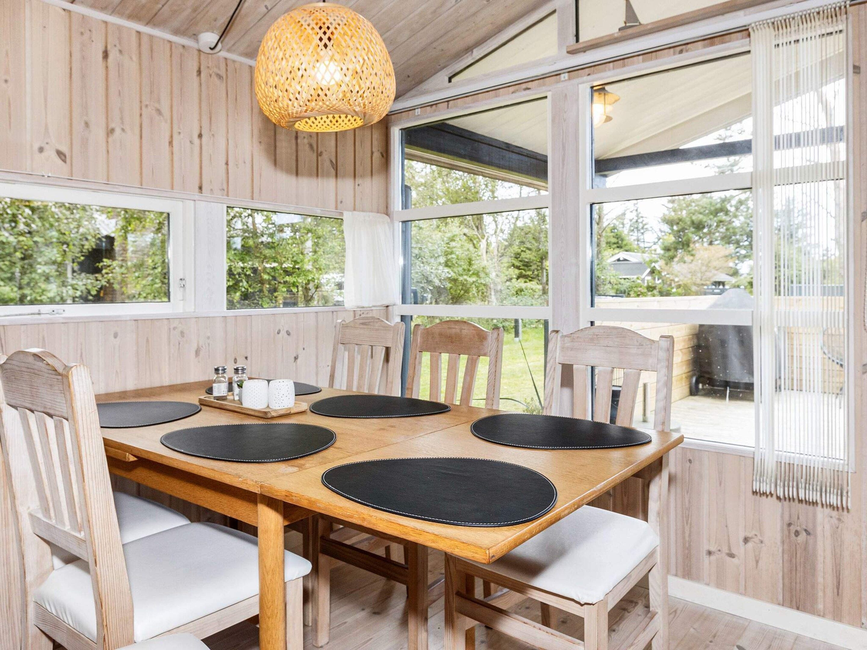 A wooden dining table with six chairs, a woven light fixture, and large windows with a view of greenery.
