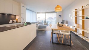 Een moderne keuken met een centraal eiland, een eethoek met een houten tafel en stoelen, en een woonruimte met een bank en een groot raam.