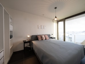 Une chambre moderne avec un grand lit, une table de chevet, une lampe fixée au mur et une vue sur la ville grâce à de grandes fenêtres.