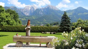 A wooden bench with a sculpture in a garden with mountains in the background. - Konigssee - Reich Family (Schoenau am Koenigssee)