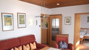A room with a red sofa, a wooden table, and a bed with a wooden headboard. There are two framed pictures on the wall and a door with frosted glass. - Lakeside Comfort in Fussen (Fuessen)