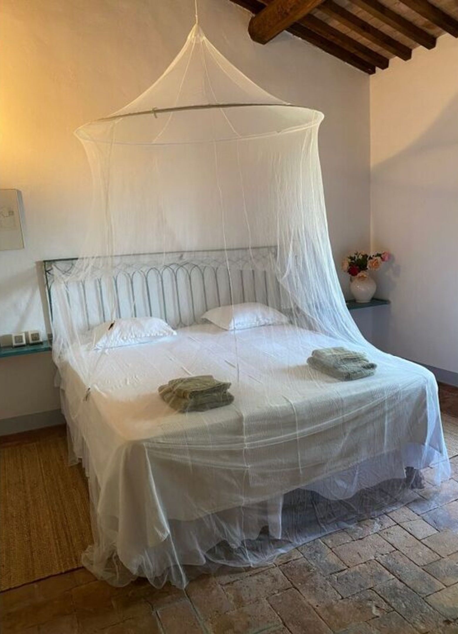 Een bed met een witte hemel, een metalen hoofdbord en een klein nachtkastje met een bloemenvaas.