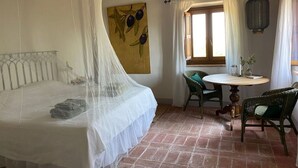 Un dormitori amb un llit sota una mosquiterola, una taula de dinar amb caires, una finestra amb cortines i un llum de llanternatge.
