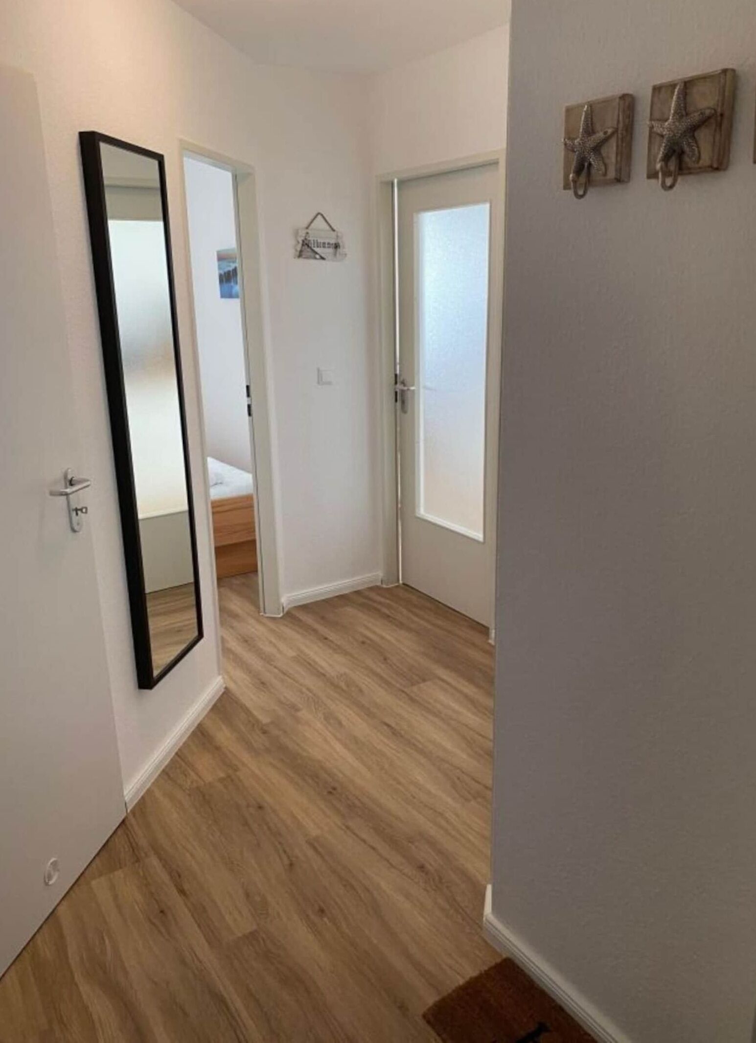 Ein Flur mit Holzboden, weißen Wänden und einer Tür zu einem Schlafzimmer. Zwei Wandschmuckstücke in Form von Meeressternen sind angebracht.