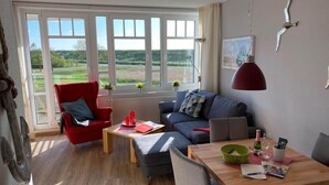 Un moderno salón con un sillón rojo, un sofá gris y una mesa de comedor de madera. Amplias ventanas ofrecen vistas al exterior.