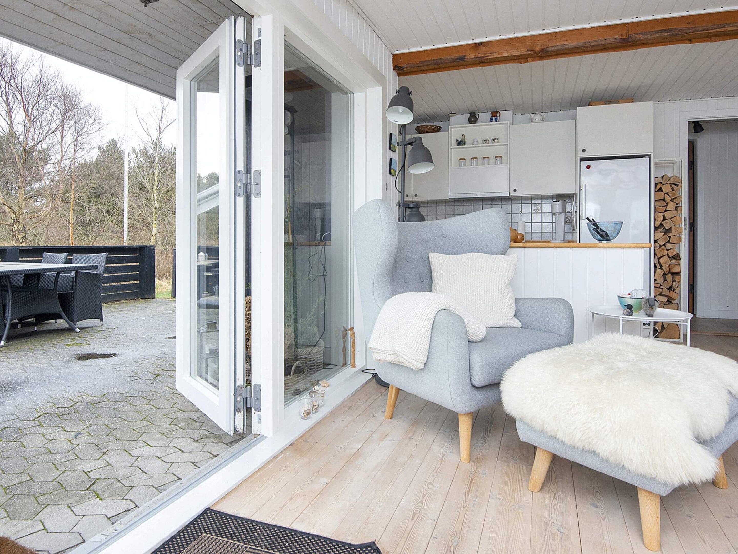 Un intérieur moderne avec un fauteuil gris, un pouf blanc et moelleux, et un coin cuisine doté de placards et d’un réfrigérateur.