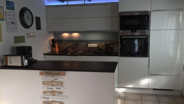 A modern kitchen with white cabinets, a black countertop, and a built-in oven. There are wall decorations and a sign with directional phrases. - Fischerhaus by the sea (Bredene)